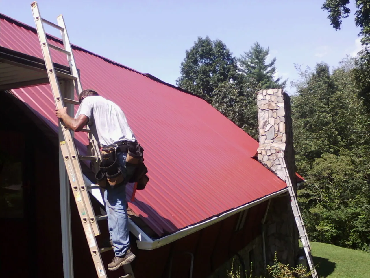 Roofer on a ladder beside a red metal roof and stone chimney in Medicine Bow
